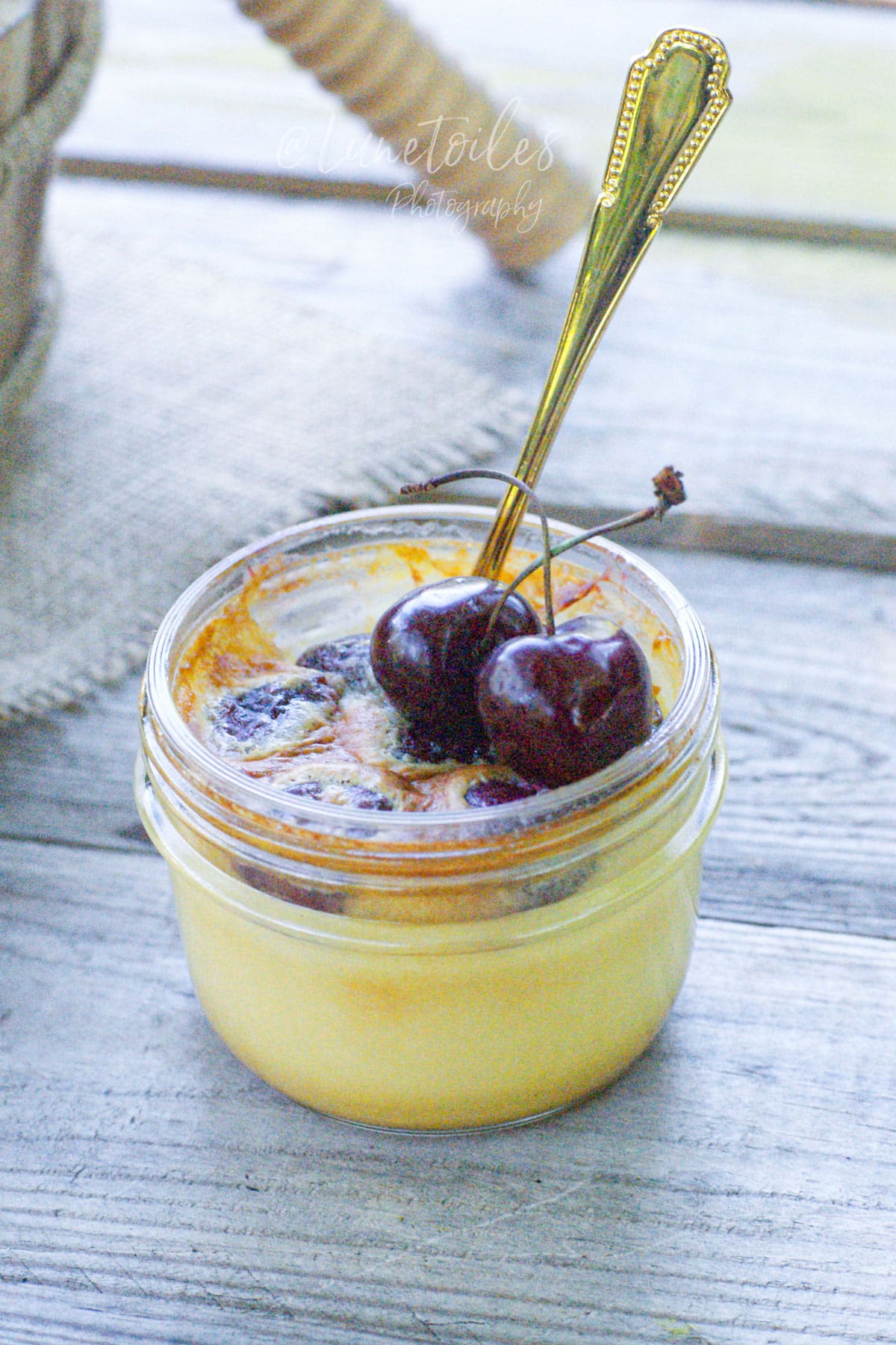 Flan aux cerises en petit pot, garni de cerises fraîches et servi avec une cuillère dorée sur une table en bois clair.