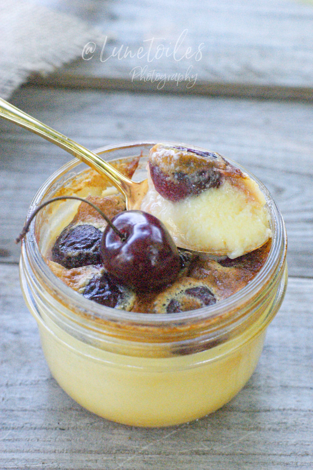Flan aux cerises en petit pot avec une cuillère dorée, montrant une texture crémeuse et des cerises fondantes après cuisson.