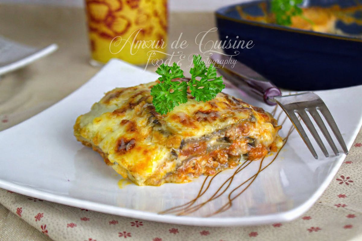 Portion de lasagnes aux aubergines gratinées, servie sur une assiette blanche et décorée de persil frais, avec une fourchette posée à côté.