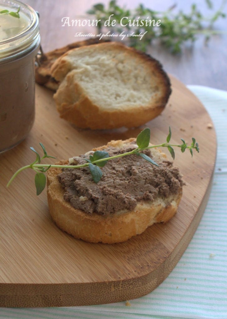 Toasted bread spread with chicken liver terrine, topped with a sprig of thyme, on a wooden board with a golden toast slice and a terrine jar blurred in the background.