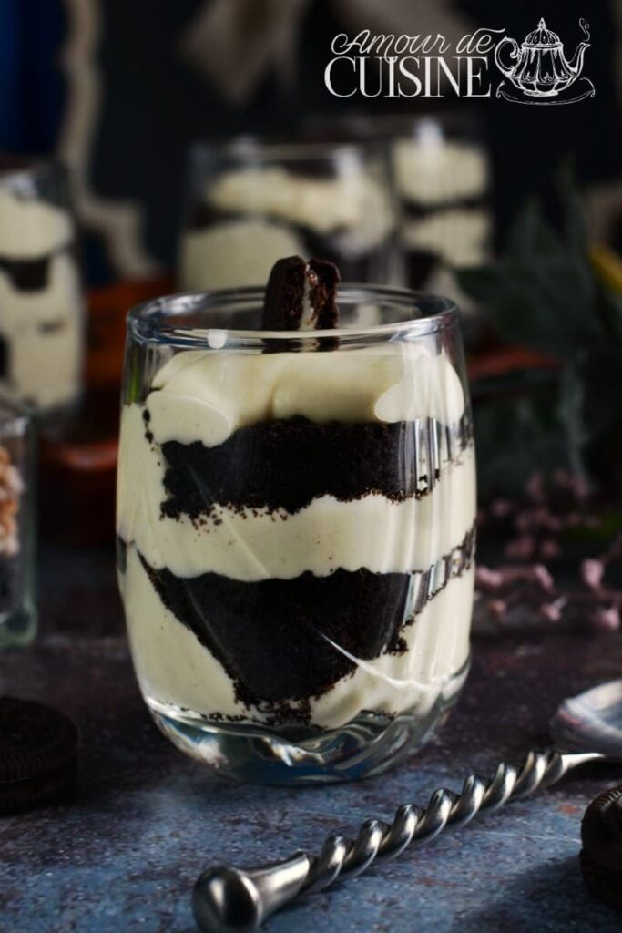 Close-up of an Oreo tiramisu served in a clear glass, showing rich layers of crushed Oreo cookies and mascarpone cream, topped with an Oreo cookie piece, on a dark textured surface with a spoon nearby.