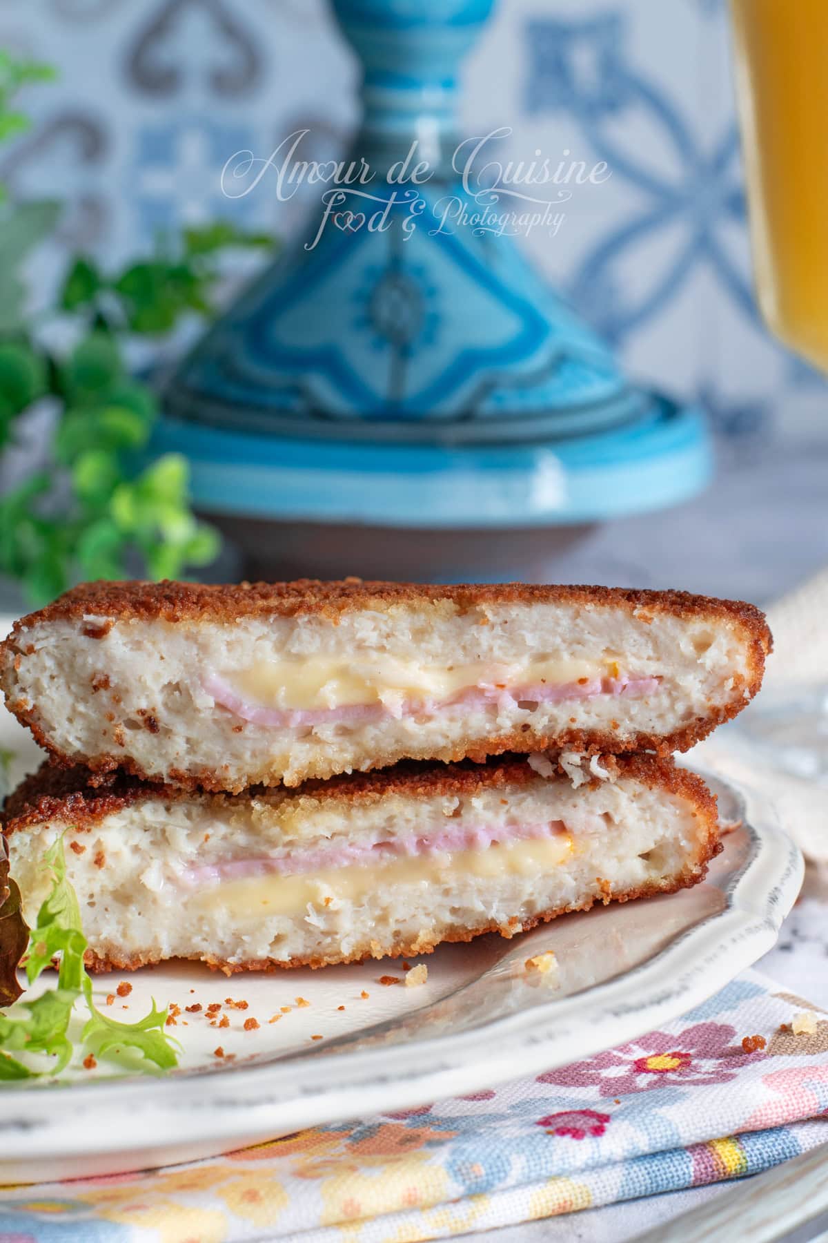 Two homemade cordon bleu cutlets cut in half and stacked, with a golden crispy breadcrumb coating and a gooey melted cheese and turkey ham center, served on a plate with a few salad leaves and a blurred blue tagine in the background.