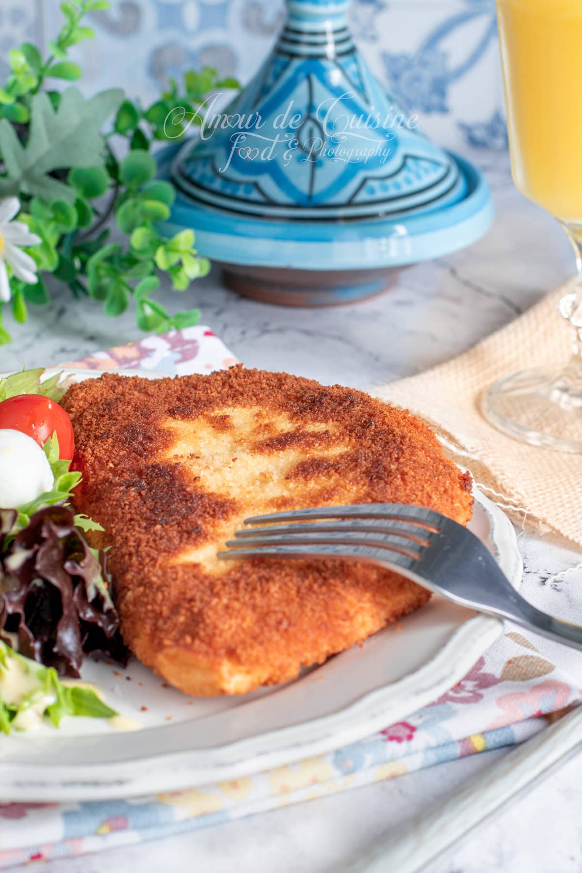 Golden homemade breaded cordon bleu served on a plate with a small mixed salad and a cherry tomato, a fork resting on top, with a blurred blue tagine in the background and a glass of juice on the side.