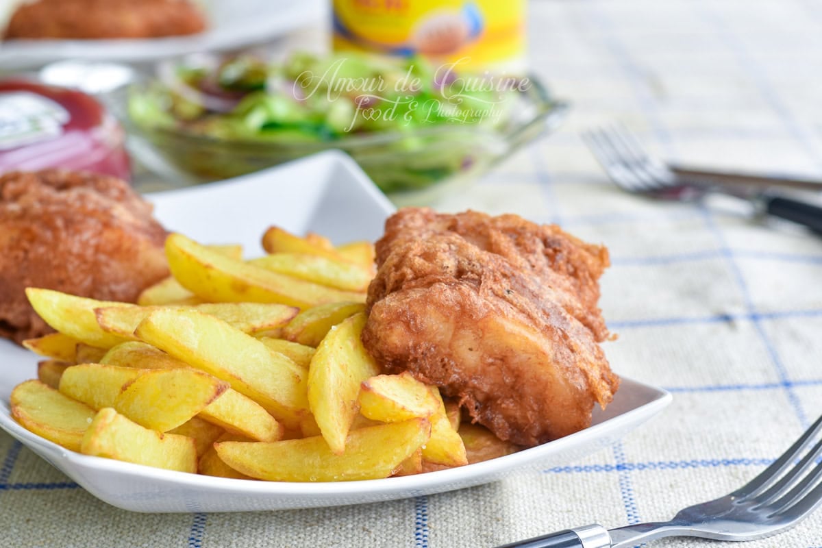 Assiette de fish and chips maison avec frites épaisses dorées et morceau de poisson frit en pâte croustillante, photographiée de côté sur une nappe à carreaux, avec salade et condiments floutés à l’arrière-plan.