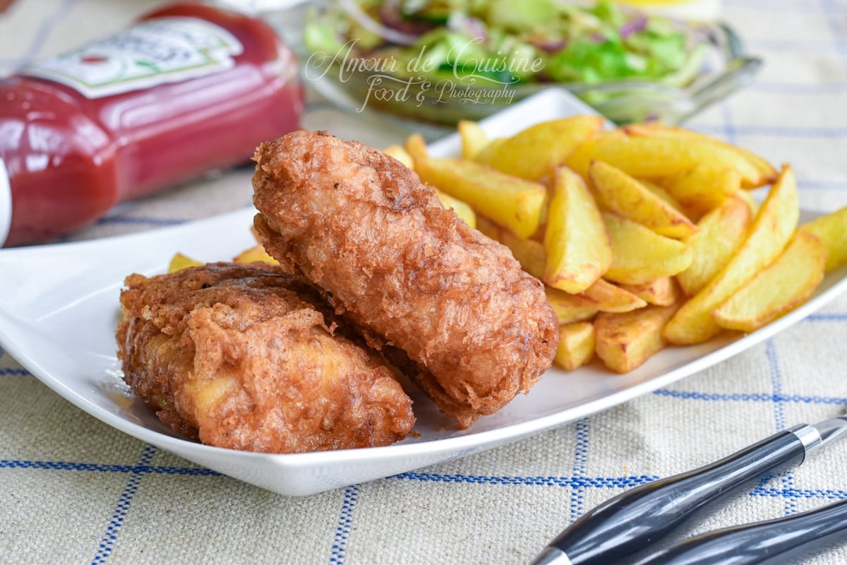Gros plan d’un fish and chips maison : deux morceaux de poisson frit en pâte dorée et des frites épaisses croustillantes dans une assiette blanche, avec ketchup et salade floutés à l’arrière-plan.