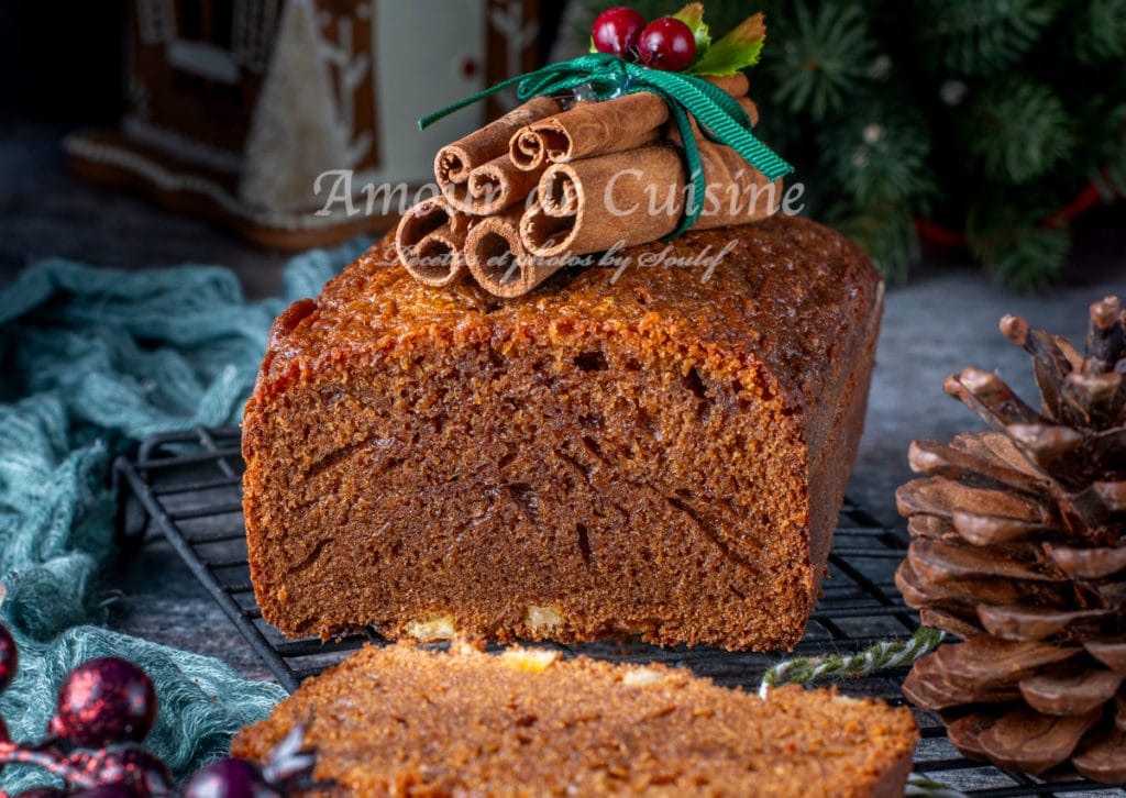 Pain d’épice moelleux au miel décoré de bâtons de cannelle, présenté dans une ambiance de Noël avec sapin et pomme de pin.