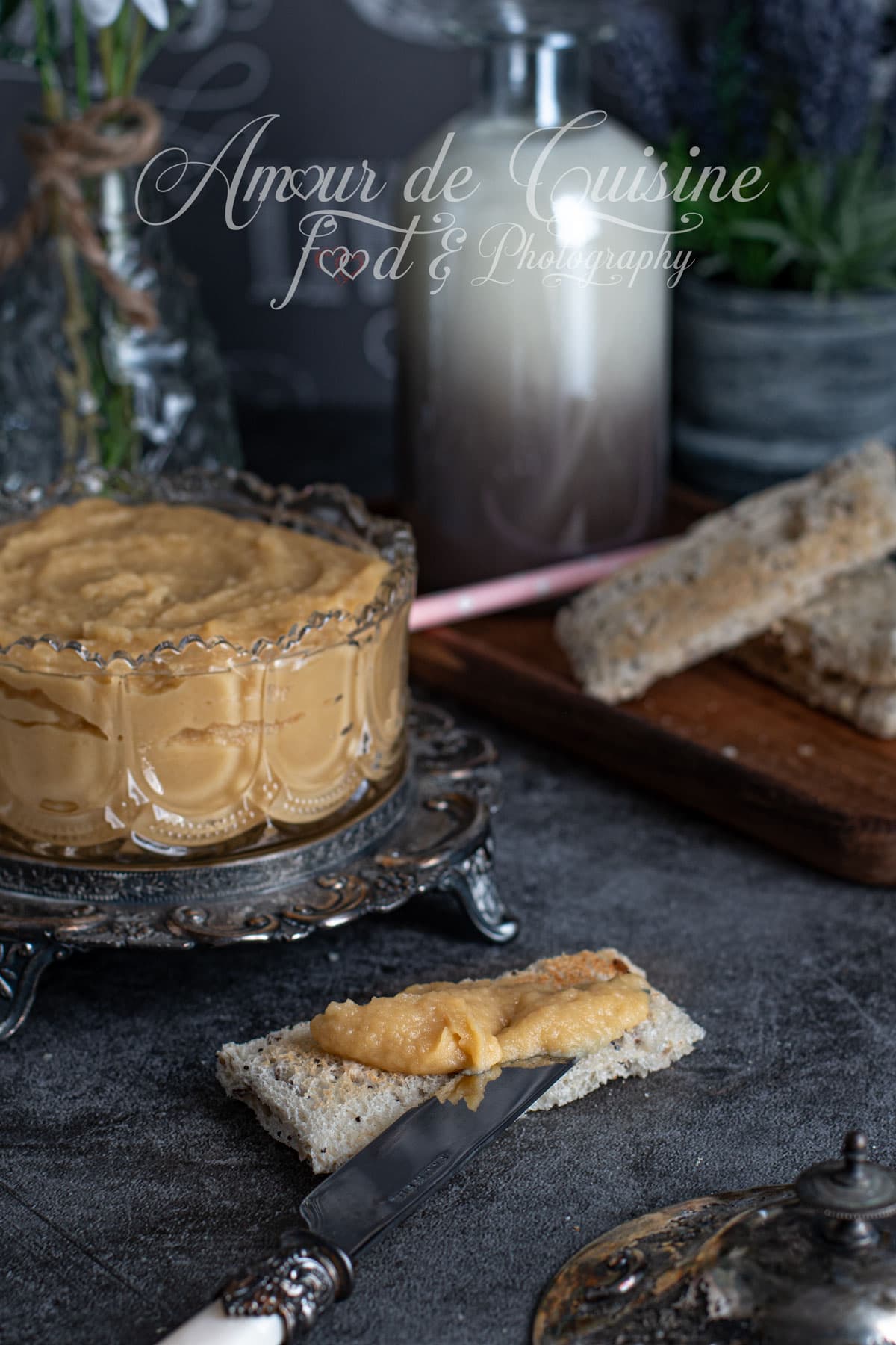 Creamy homemade apple butter served in a glass bowl on a silver tray, with a slice of toast spread in the foreground and a rustic background setting.