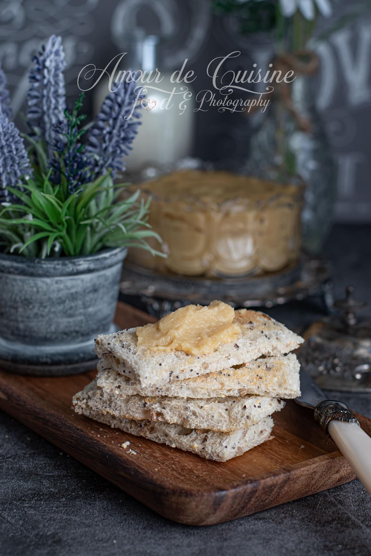 Stacked slices of multigrain bread on a wooden board, topped with a spoonful of creamy apple butter, with a decorative plant and a blurred bowl of apple butter in the background.