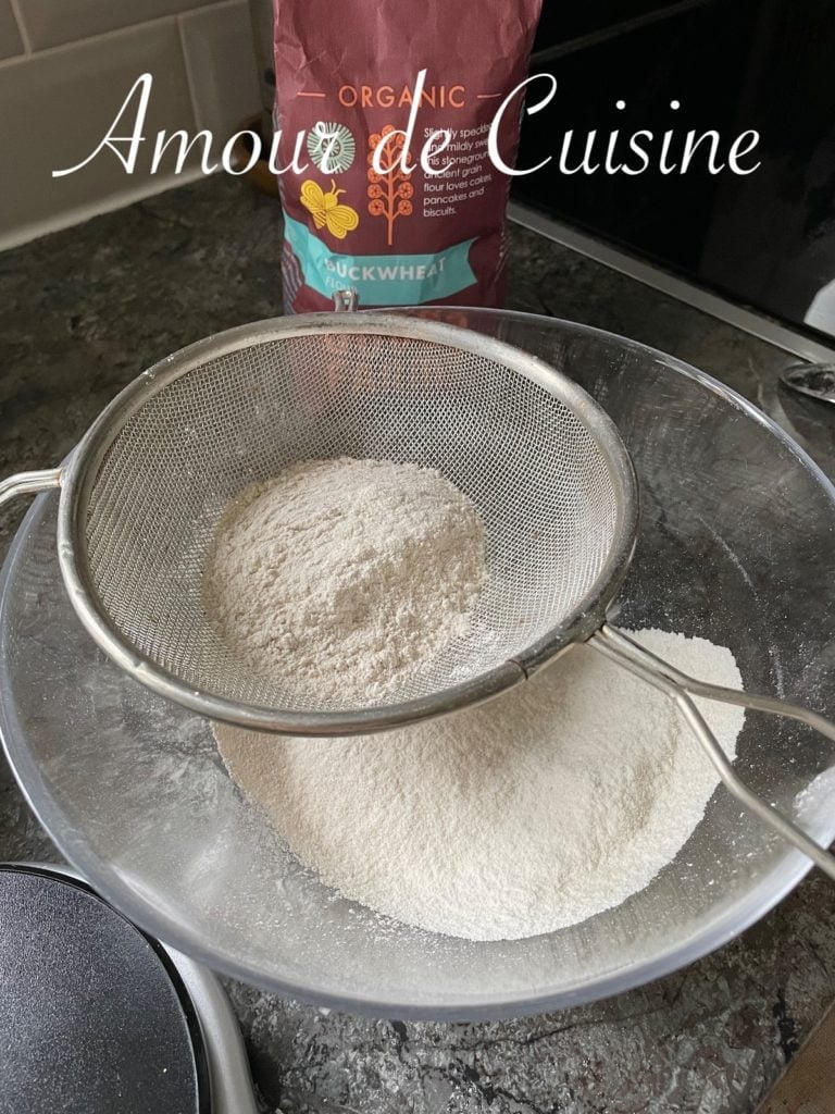 Buckwheat flour being sifted through a metal sieve into a large mixing bowl on a kitchen countertop, with a bag of organic buckwheat flour in the background.