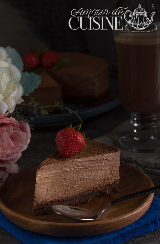 A slice of rich chocolate mousse cake topped with a fresh strawberry, served on a wooden plate with a spoon.