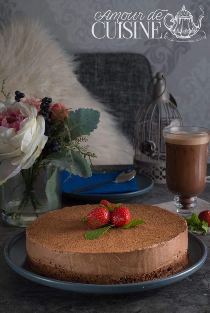 A decadent gluten-free chocolate mousse cake topped with fresh strawberries and mint leaves, elegantly presented on a dark blue plate. The scene is set with a vase of flowers, a cup of hot chocolate, and a cozy background featuring a fluffy pillow and decorative birdcage, creating a warm and inviting ambiance. The image is branded with the “Amour de Cuisine” logo at the top