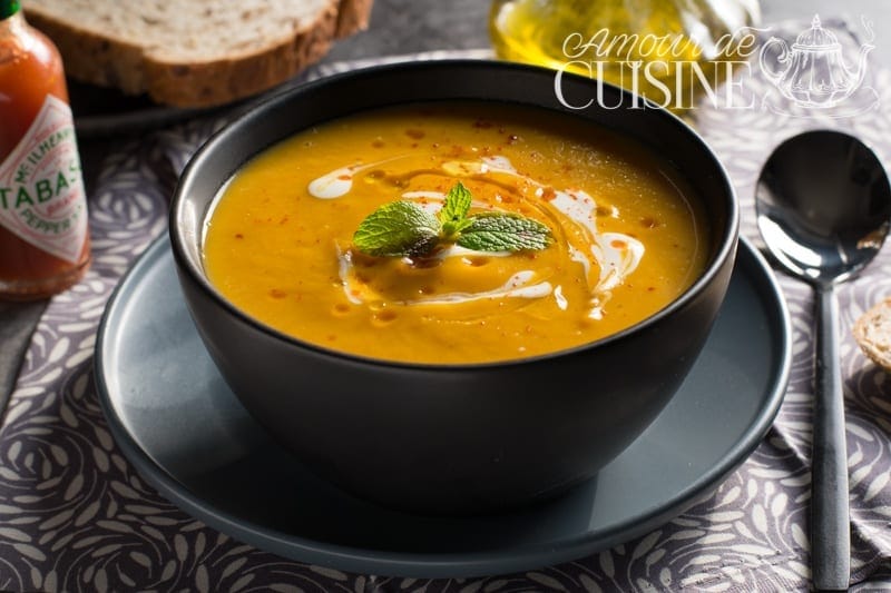 Bowl of creamy butternut squash soup, topped with fresh cream and mint leaves, served on a plate with a spoon, accompanied by bread and olive oil.