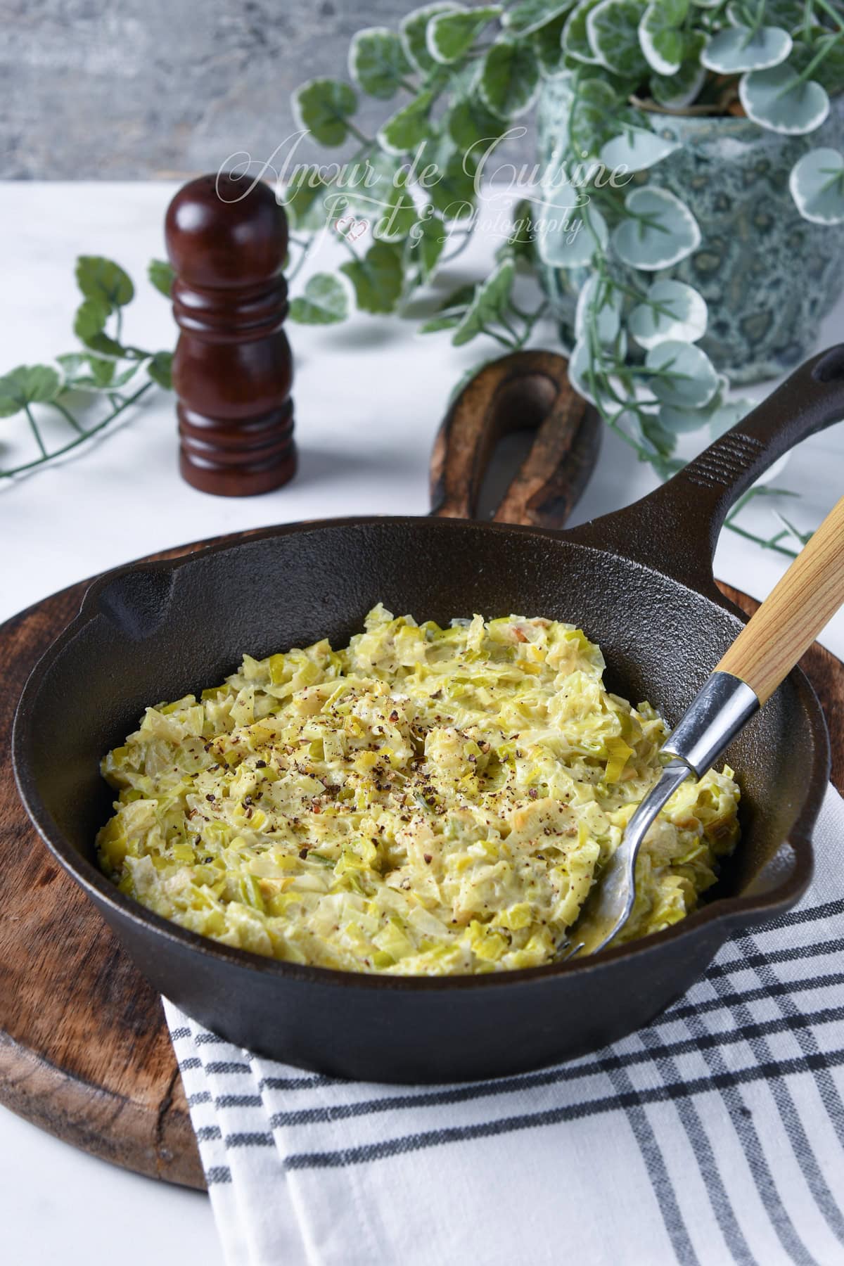 Creamy leek fondue served in a cast iron skillet, sprinkled with black pepper, with a wooden spoon, placed on a rustic wooden board.