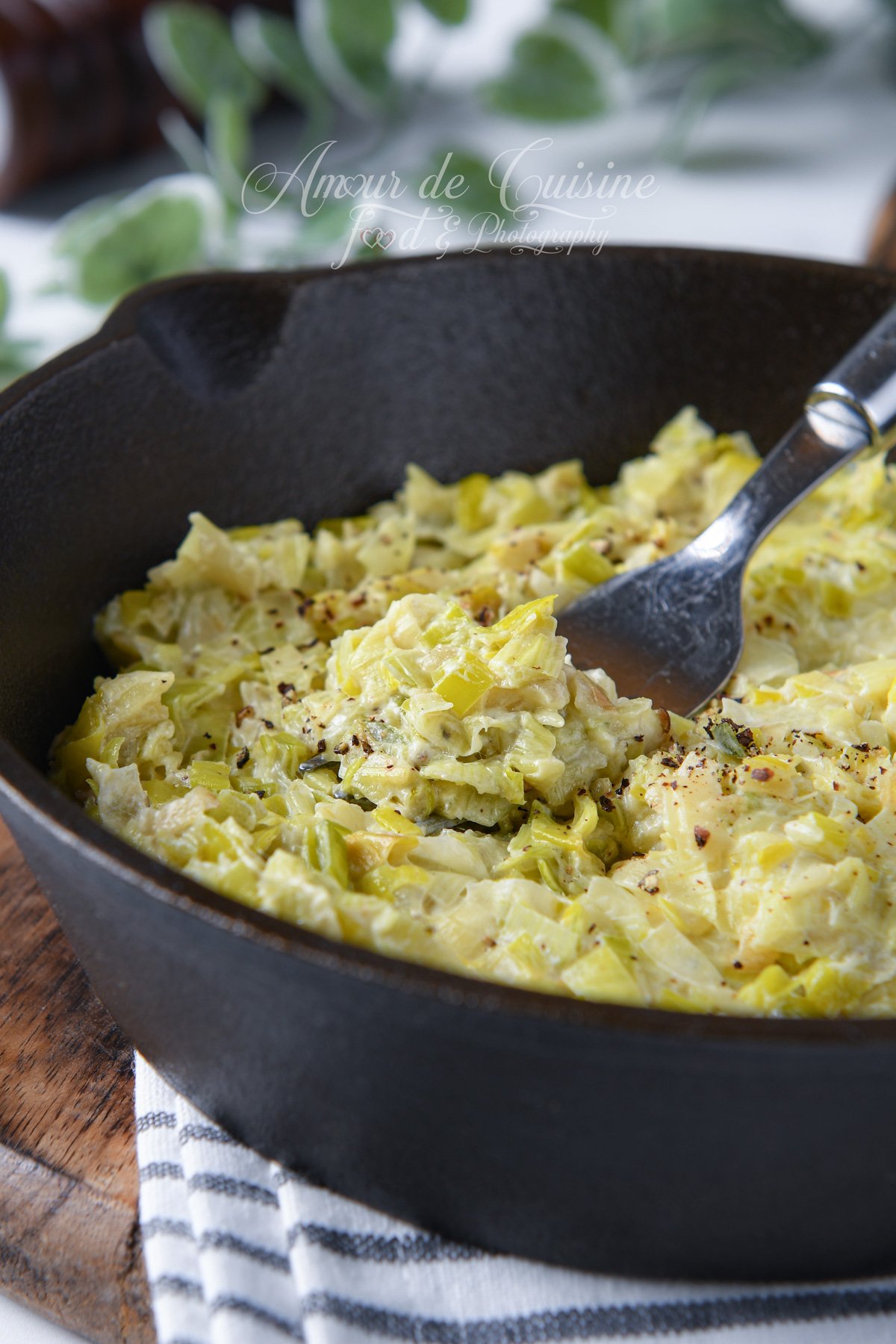 Close-up of creamy leek fondue in a cast iron skillet, with a spoon serving a portion, topped with freshly ground black pepper.