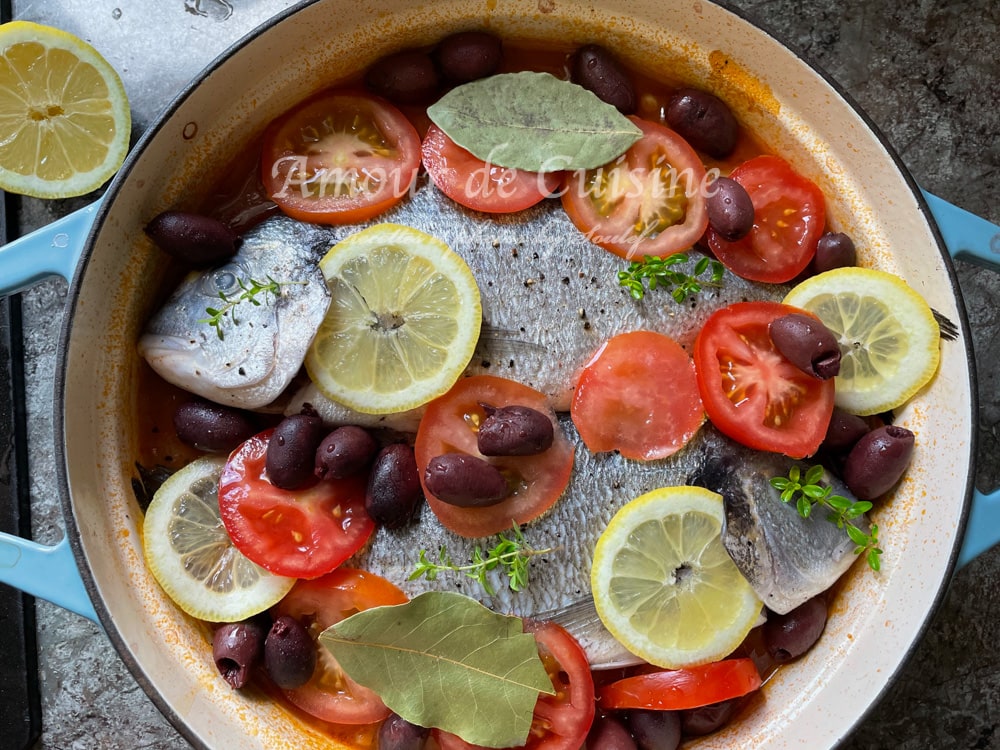 Uncooked royal dorade fish (sea bream) arranged in a baking dish over a tomato and potato base, garnished with lemon slices, fresh tomato rounds, black olives, bay leaves, and fresh herbs, ready to be baked in the oven