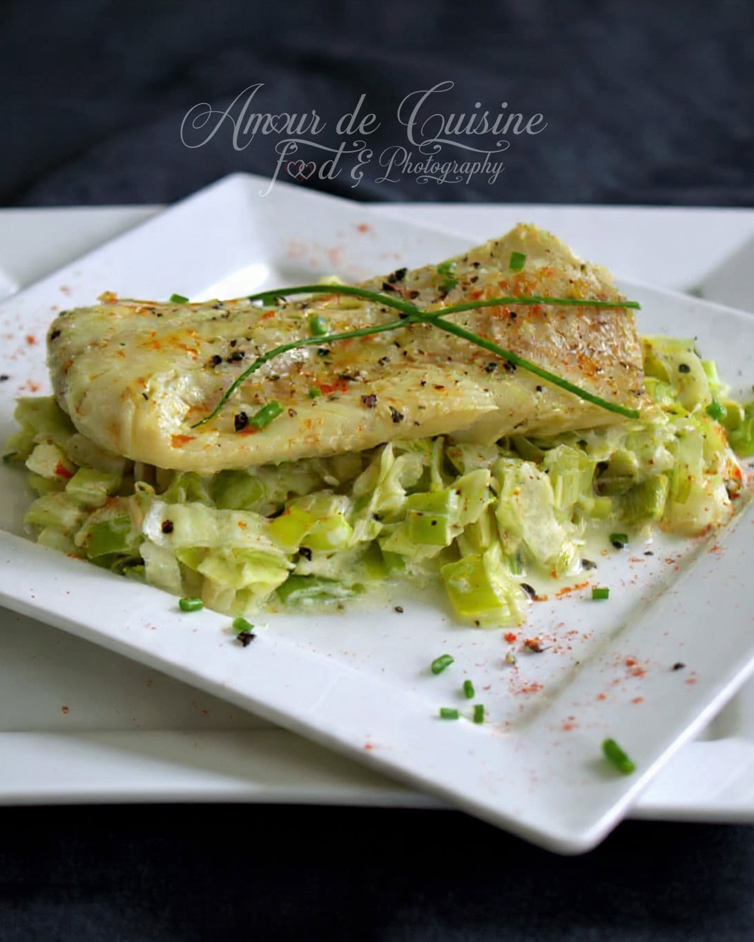 Golden, seasoned cod fillet served over a creamy leek fondue, presented on a square white plate and garnished with fresh chives and a pinch of paprika.