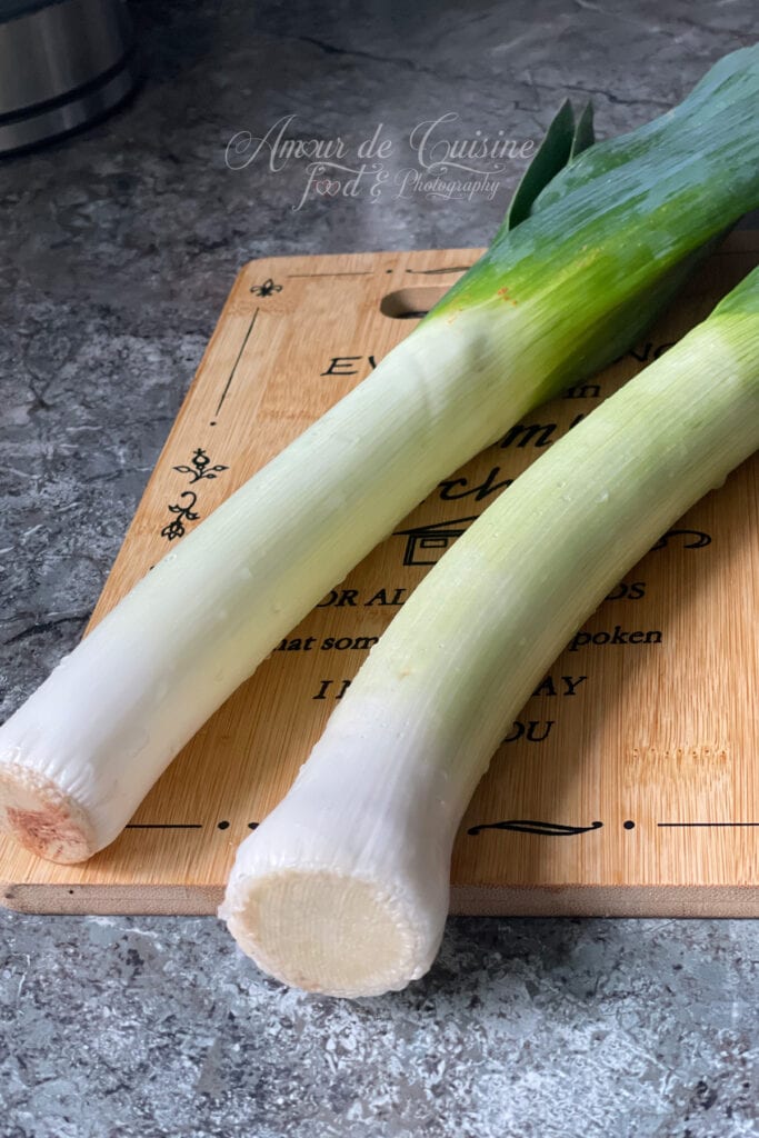 Two whole fresh leeks placed on a wooden cutting board, showing the white and light green parts, ready to be prepared.