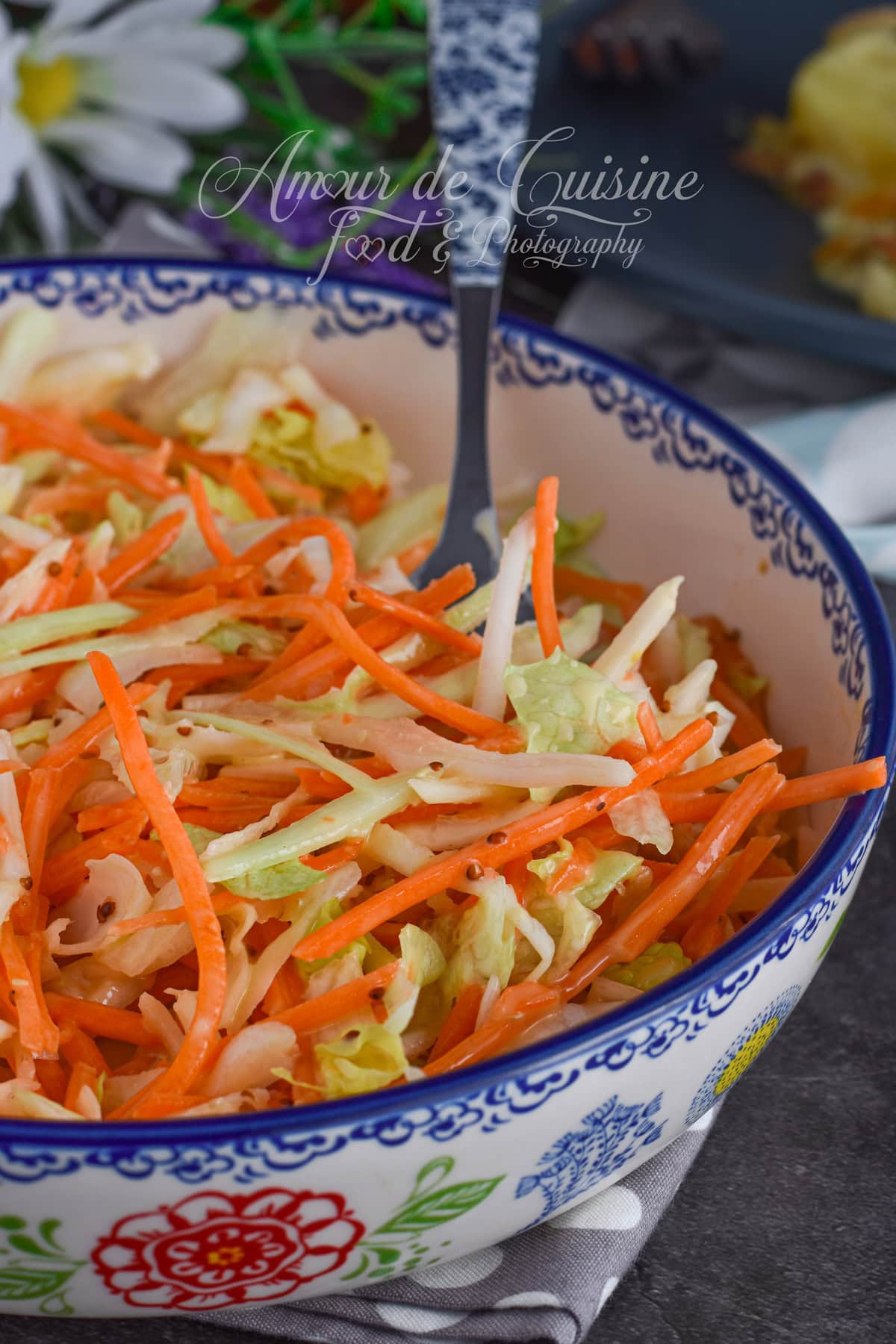 Decorative bowl filled with homemade coleslaw made of shredded white cabbage and grated carrots, set on a wooden board, with a fork in the salad and a decorative plant in the background.