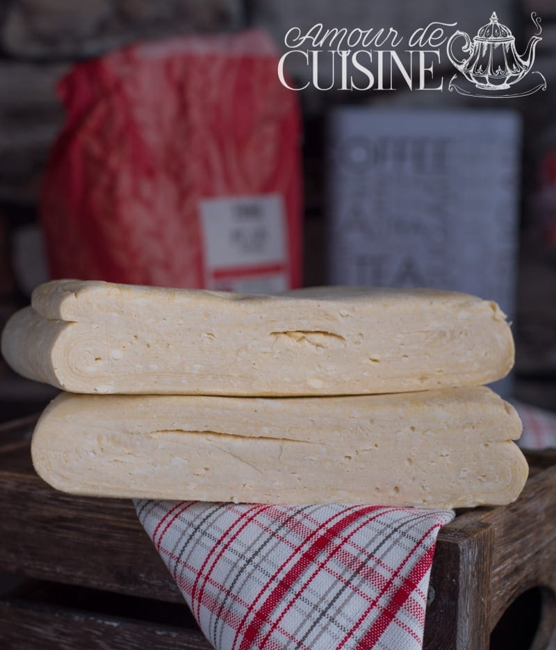 Two blocks of homemade butter puff pastry dough stacked on a wooden tray, with a red-and-grey check tea towel and a softly blurred kitchen background.