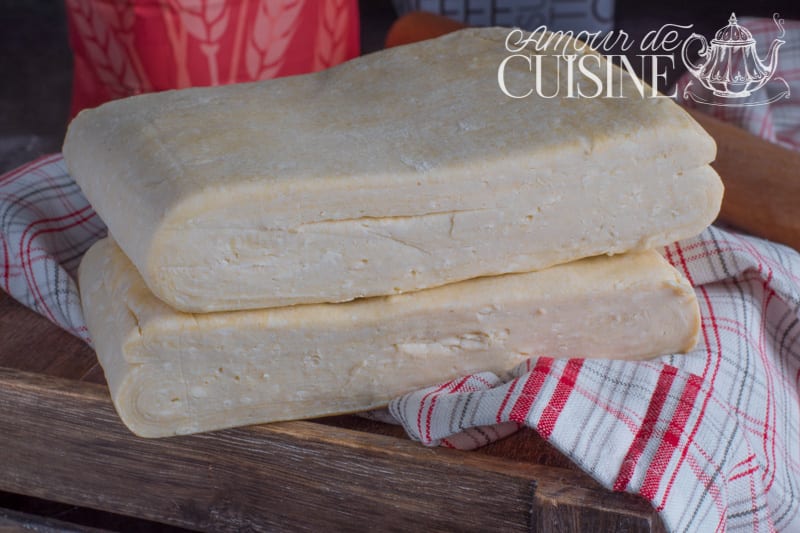 Two blocks of homemade butter puff pastry dough stacked on a wooden board, resting on a red-and-white check tea towel.