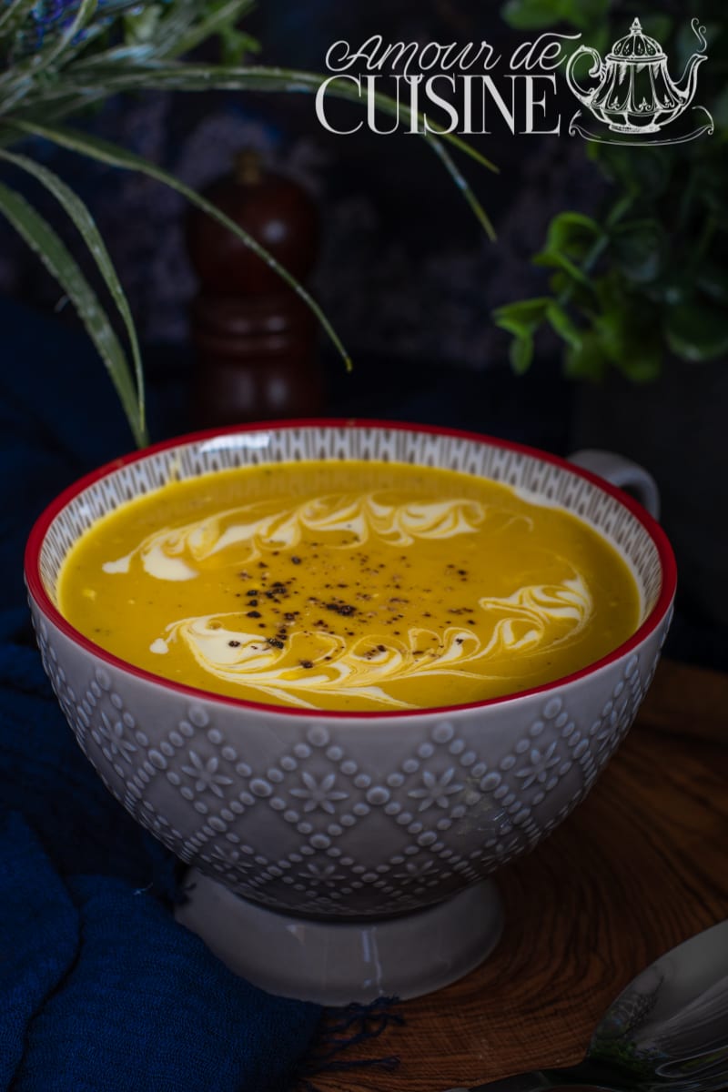 Close-up of a grey bowl of sweet potato and zucchini curry soup, topped with a cream swirl and black pepper, set on a wooden board with a blue cloth and greenery in the background.