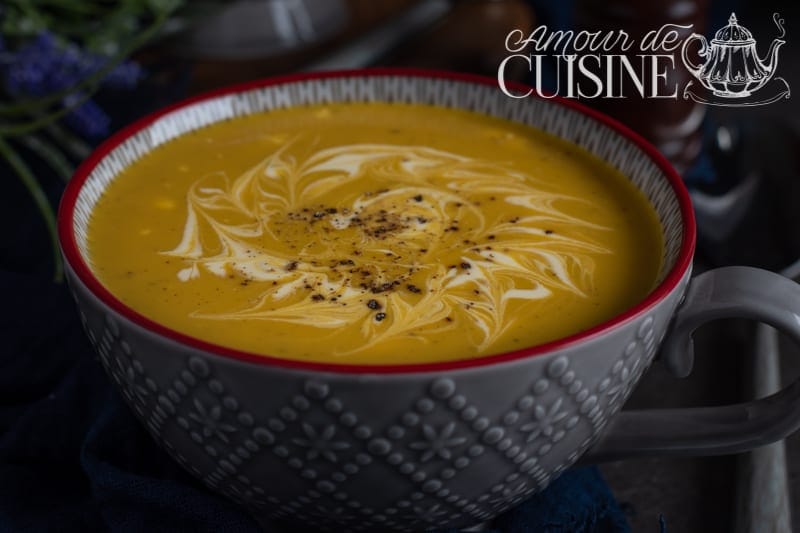 Grey bowl of sweet potato and zucchini curry soup, topped with a cream swirl and a sprinkle of black pepper, set on a blue cloth, with the Amour de Cuisine logo at the top.