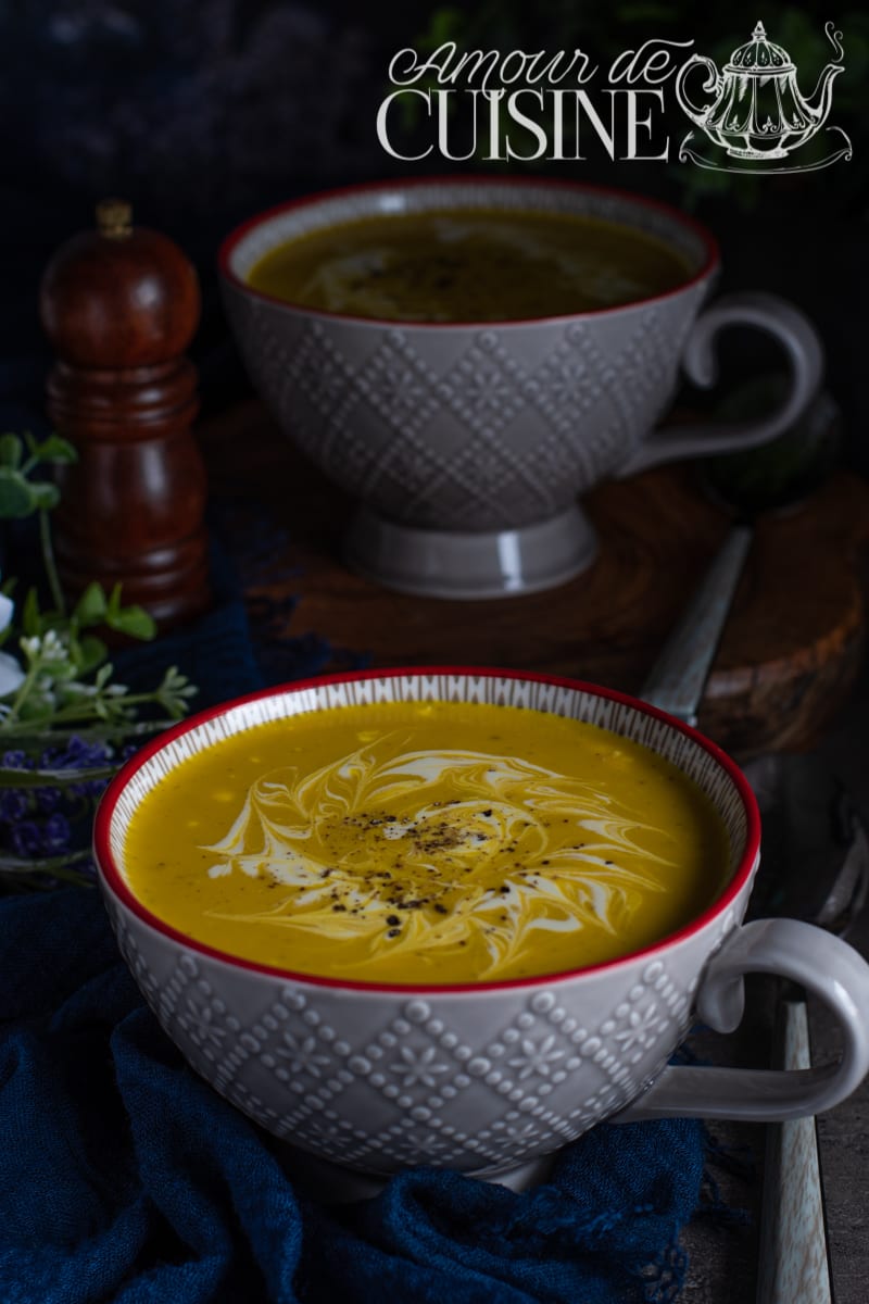 Two large grey cups of sweet potato and zucchini curry soup, topped with a cream swirl and black pepper, styled on a dark background with a pepper mill and a blue cloth.