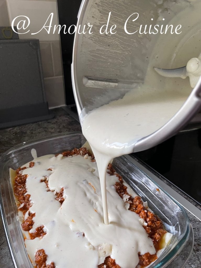 Creamy béchamel sauce being poured from a saucepan over a layer of meat sauce in a glass baking dish, preparing homemade lasagna.