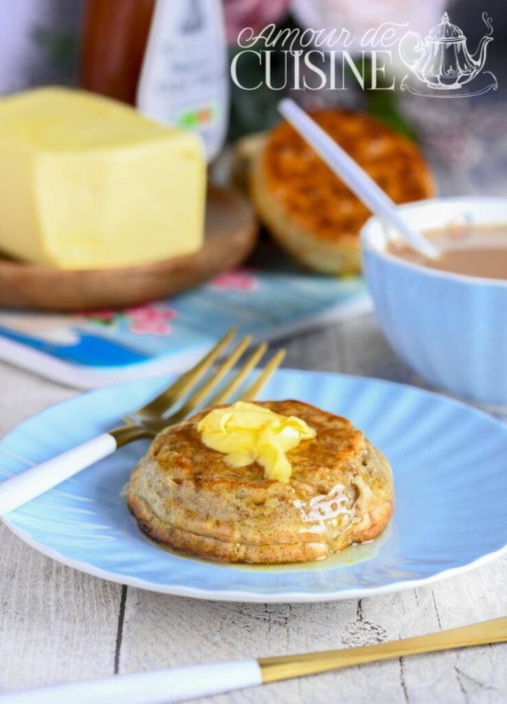 Wholemeal crumpet topped with melting butter and honey on a blue plate, with a cup of tea and butter in the background.