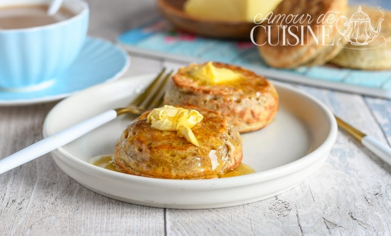 Two wholemeal crumpets topped with melting butter and honey on a white plate, with a cup of tea and extra crumpets in the background.