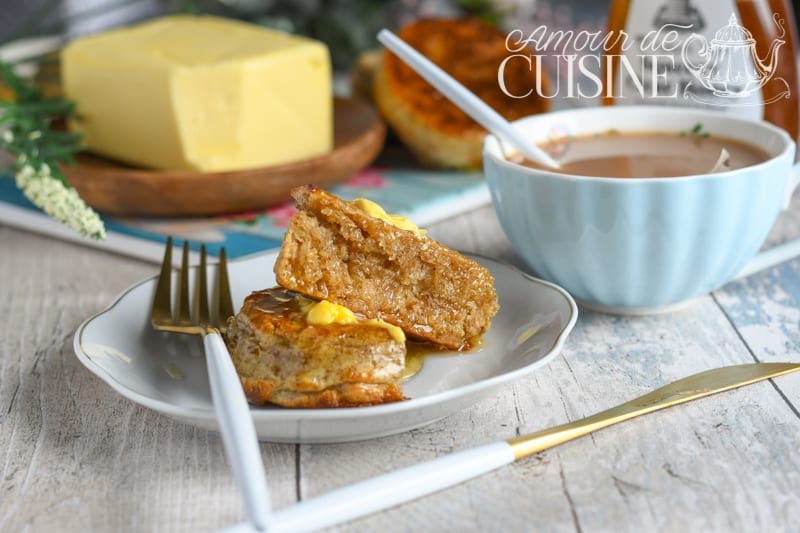 Wholemeal crumpet cut open and stacked on a small plate with melting butter and honey, served with a cup of tea and a block of butter in the background.