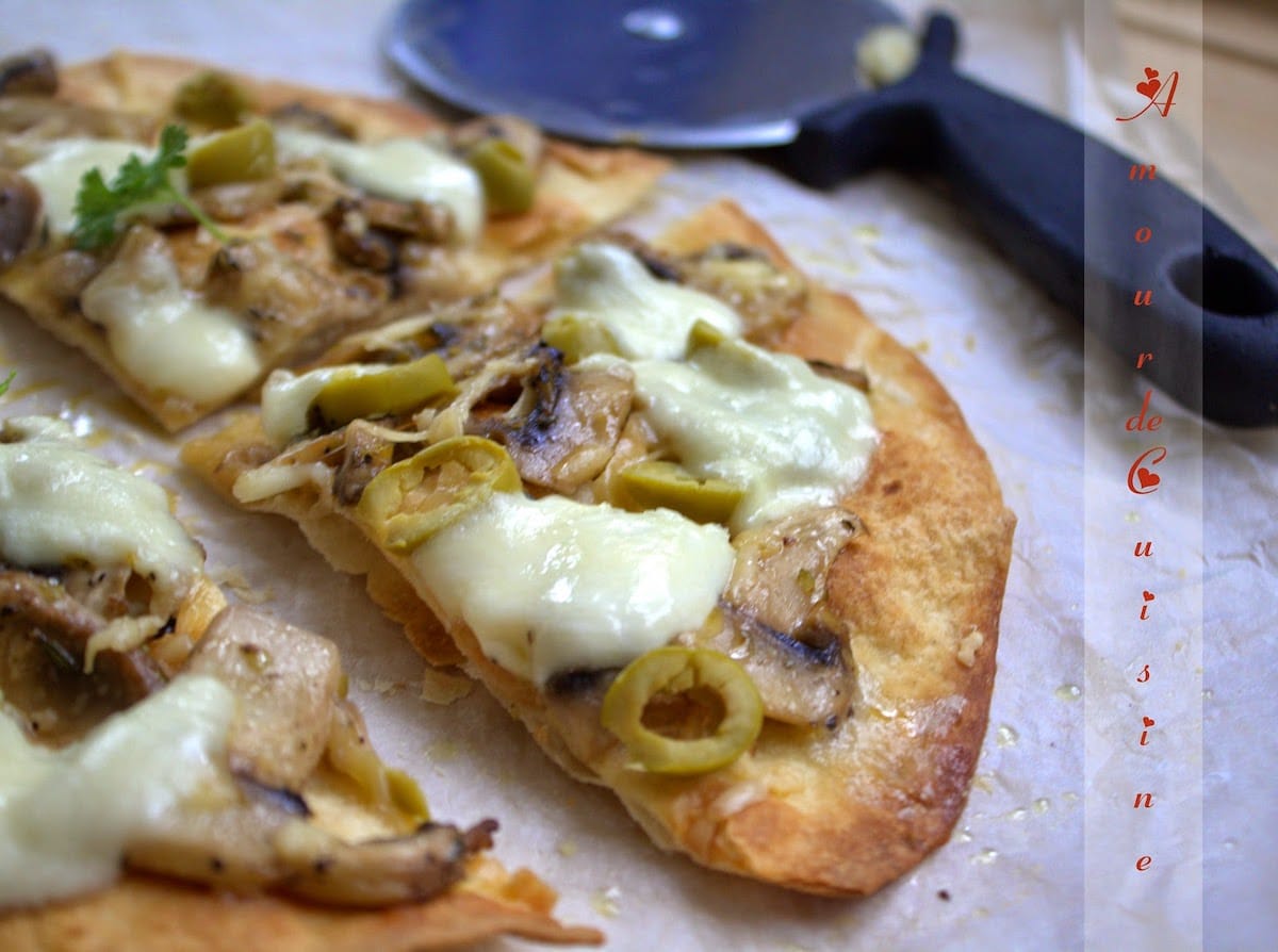 Tranches de pizza tortillas garnies de champignons, olives vertes et fromage fondu, posées sur du papier cuisson avec une roulette à pizza en arrière-plan
