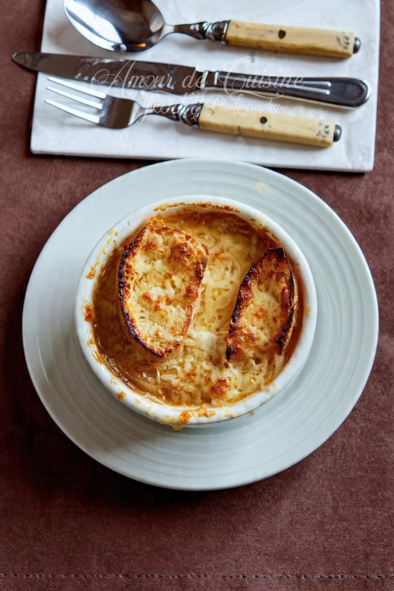 Bowl of French onion soup seen from above, served in a white ramekin with two baguette slices topped with golden cheese, placed on a white plate with cutlery in the background.