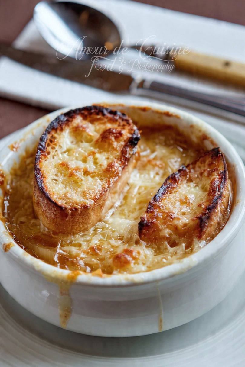 Close-up of French onion soup in a white ramekin, served with two crispy baguette slices topped with golden cheese, with a spoon and knife blurred in the background.