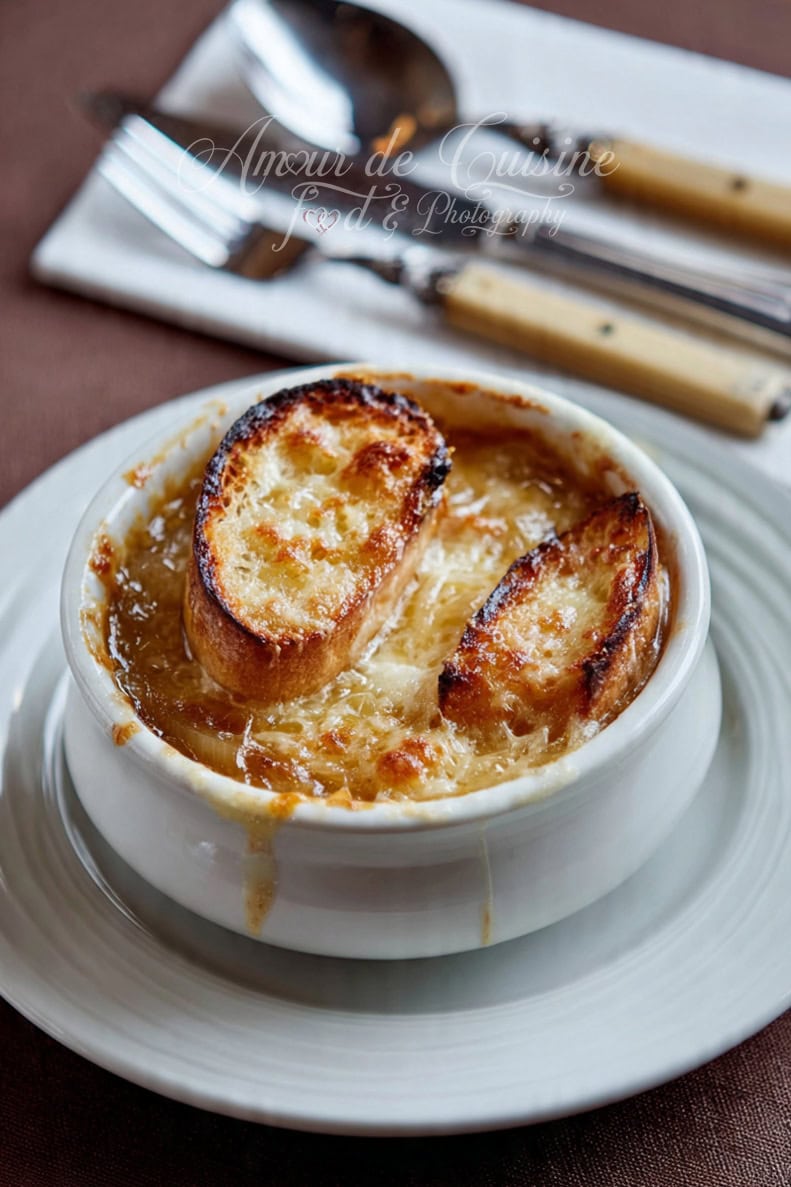 Bowl of French onion soup in a white ramekin, topped with baguette slices covered in golden, melted cheese, placed on a plate with blurred cutlery in the background.