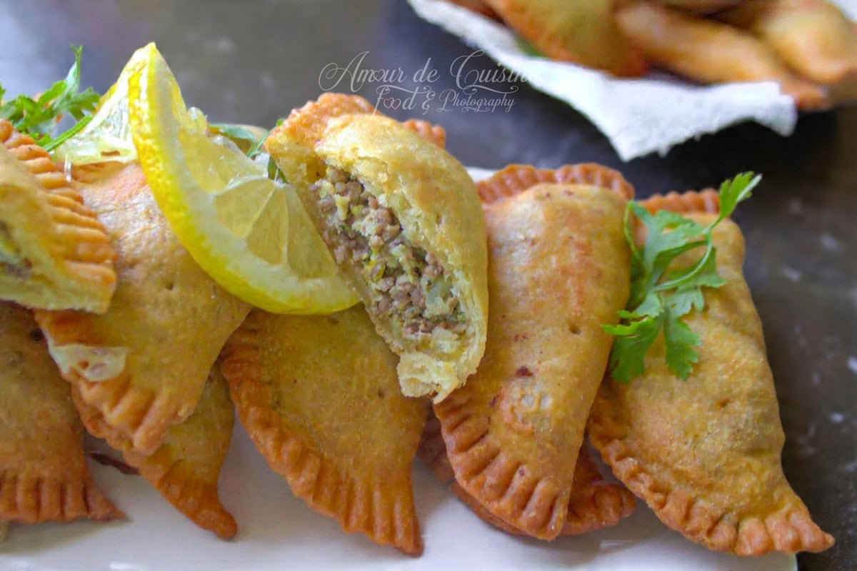 Golden Algerian empanadas (bourek laadjine) lined up on a white platter, with one piece opened to show the minced meat filling, garnished with lemon slices and parsley on a dark surface.