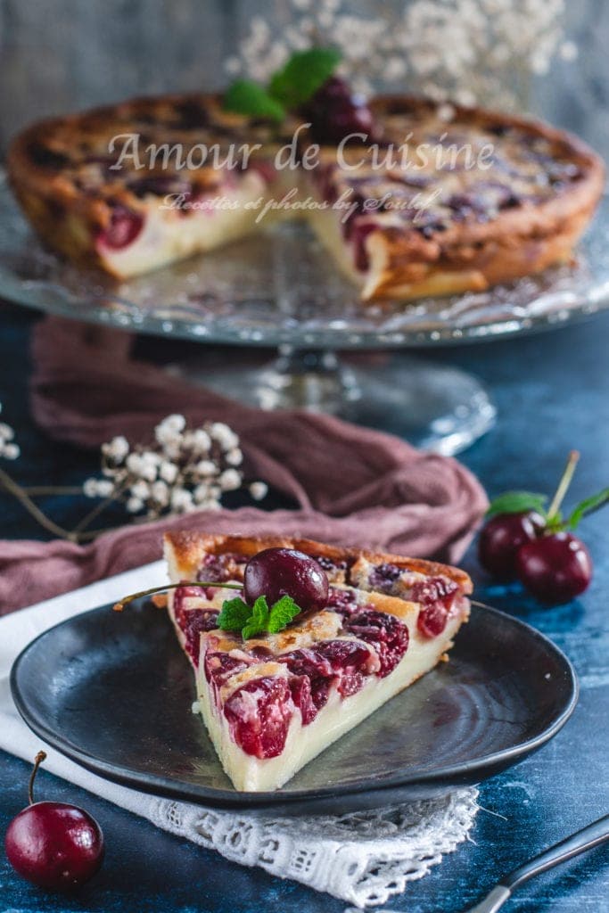 Slice of homemade French cherry clafoutis served on a black plate, topped with a fresh cherry and mint, with the full tart in the background on a glass cake stand
