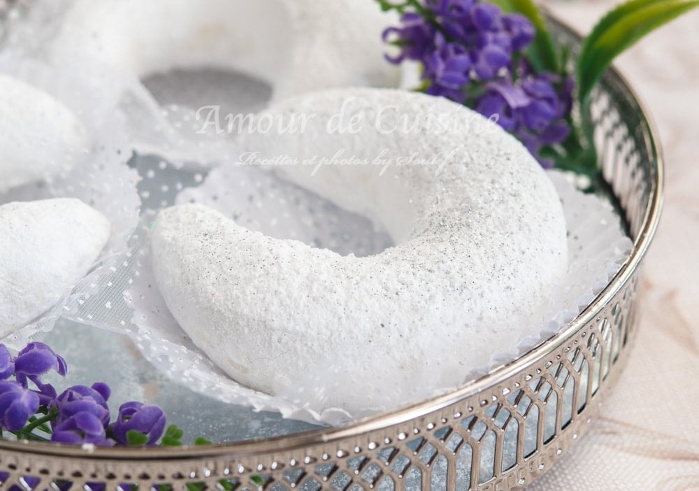 Powdered sugar–coated Algerian tcharek msaker crescent cookie served on a silver tray with a lace doily and purple flowers.
