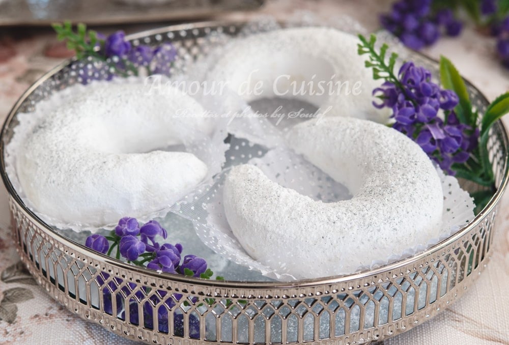 Iced sugar–coated Algerian tcharek msaker crescent cookies arranged on a silver tray with a lace doily and purple flowers.