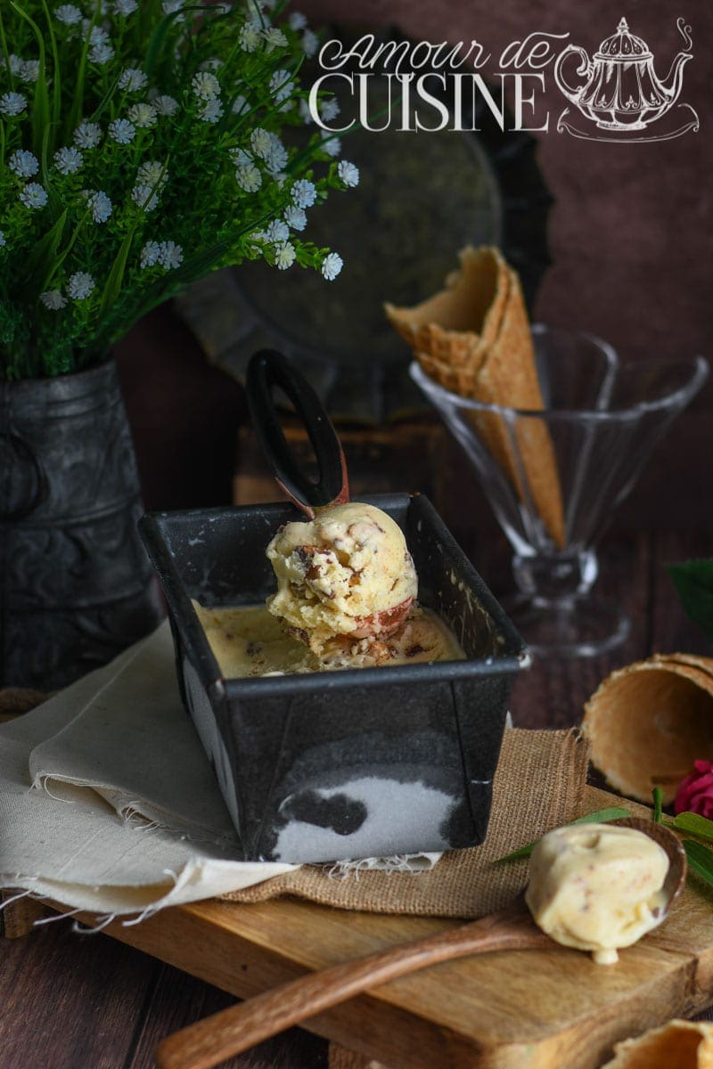 Crème glacée à la vanille maison servie dans un bac noir, avec éclats de fruits secs, cuillère à glace, cornets en arrière-plan et décor champêtre