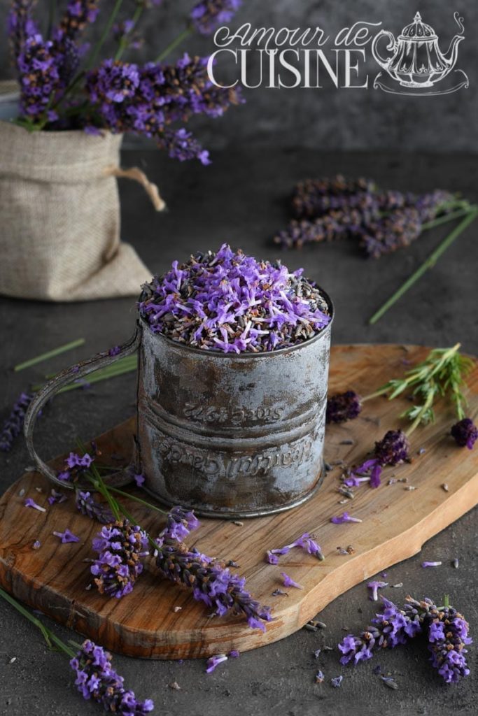 A rustic metal cup filled with dried and fresh lavender flowers, displayed on a wooden board with scattered lavender sprigs, while a burlap-wrapped bouquet sits in the background against a dark, textured surface.