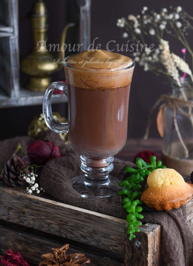 Glass of cocoa mocha coffee topped with thick dalgona foam, set on a rustic tray with a small cake and a touch of greenery.