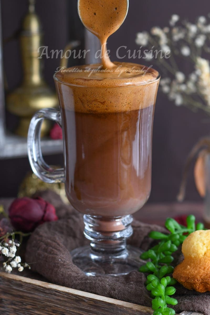 Cocoa mocha coffee in a glass, topped with thick dalgona foam being spooned on, with a rustic background setting.