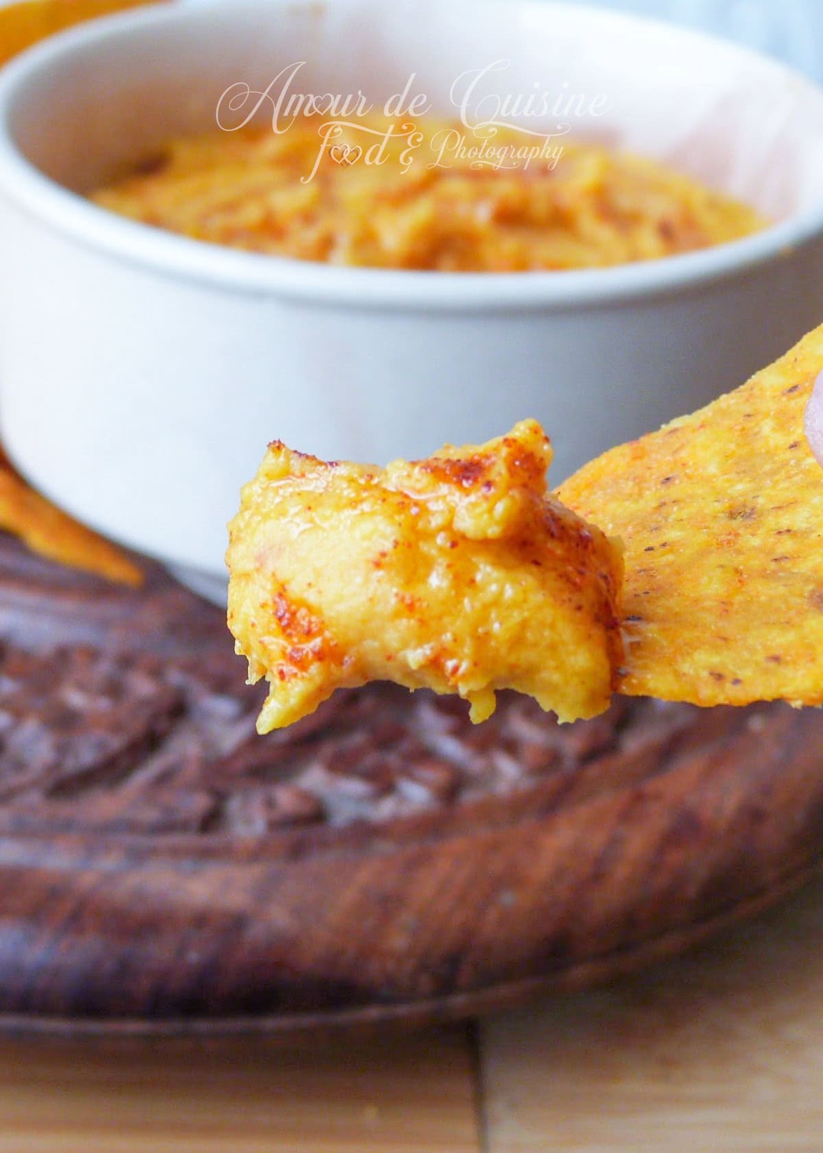 Close-up of creamy sweet potato hummus with a hint of paprika, scooped onto a crispy tortilla chip, with a bowl of hummus in the background.
