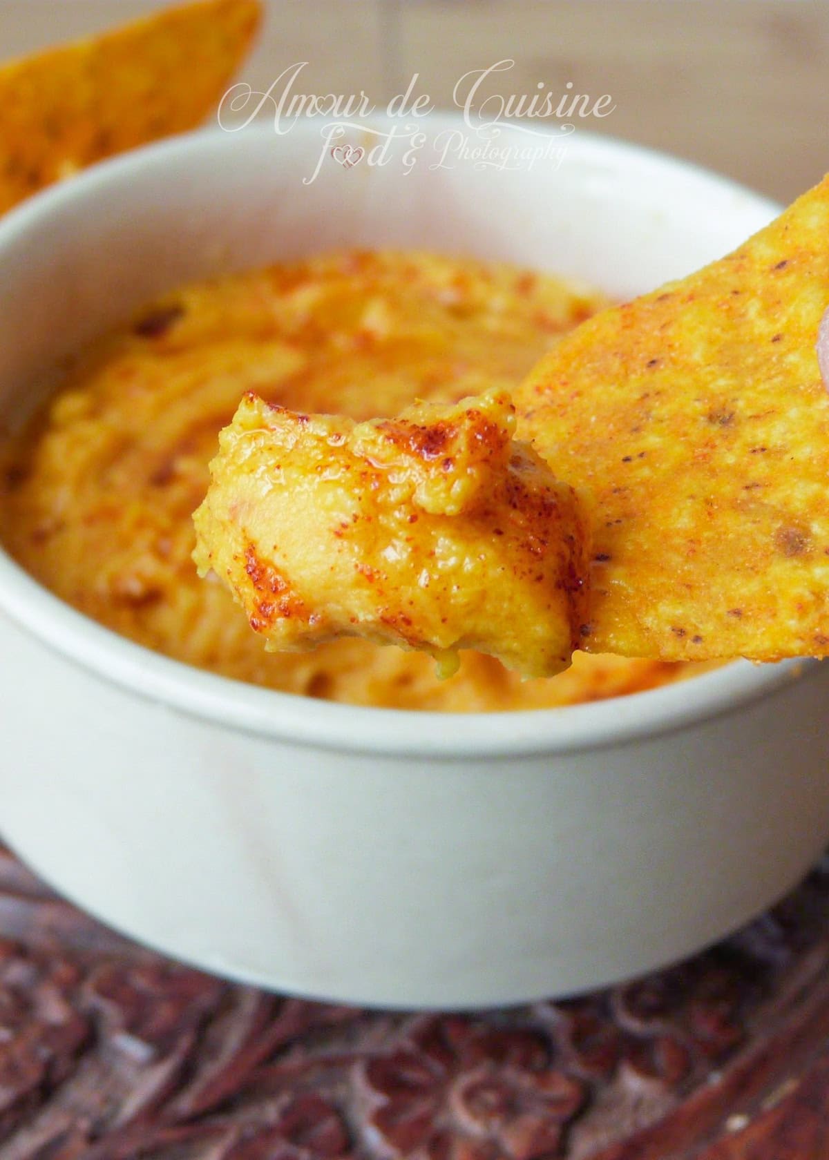 Close-up of sweet potato hummus scooped onto a seasoned tortilla chip, with a creamy bowl of hummus in the background.