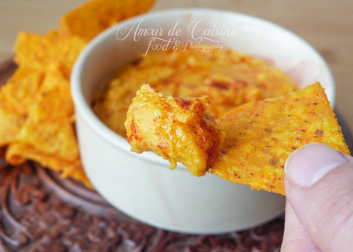 Scoop of sweet potato hummus on a spiced tortilla chip, held in hand, with a bowl of hummus and a pile of chips in the background.
