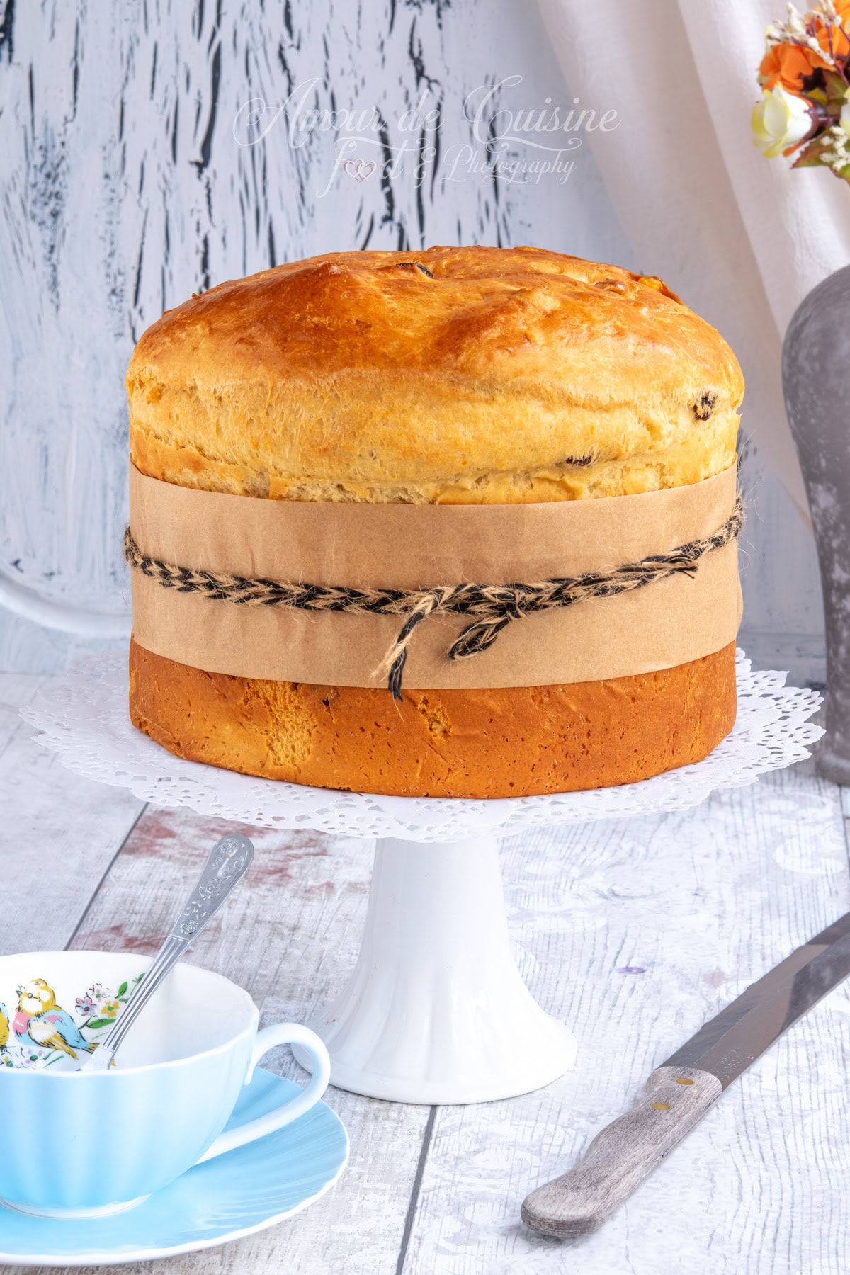 Panettone maison doré présenté entier sur un présentoir blanc, entouré d’une bande de papier kraft et d’une ficelle, avec une tasse bleue et un couteau sur la table.