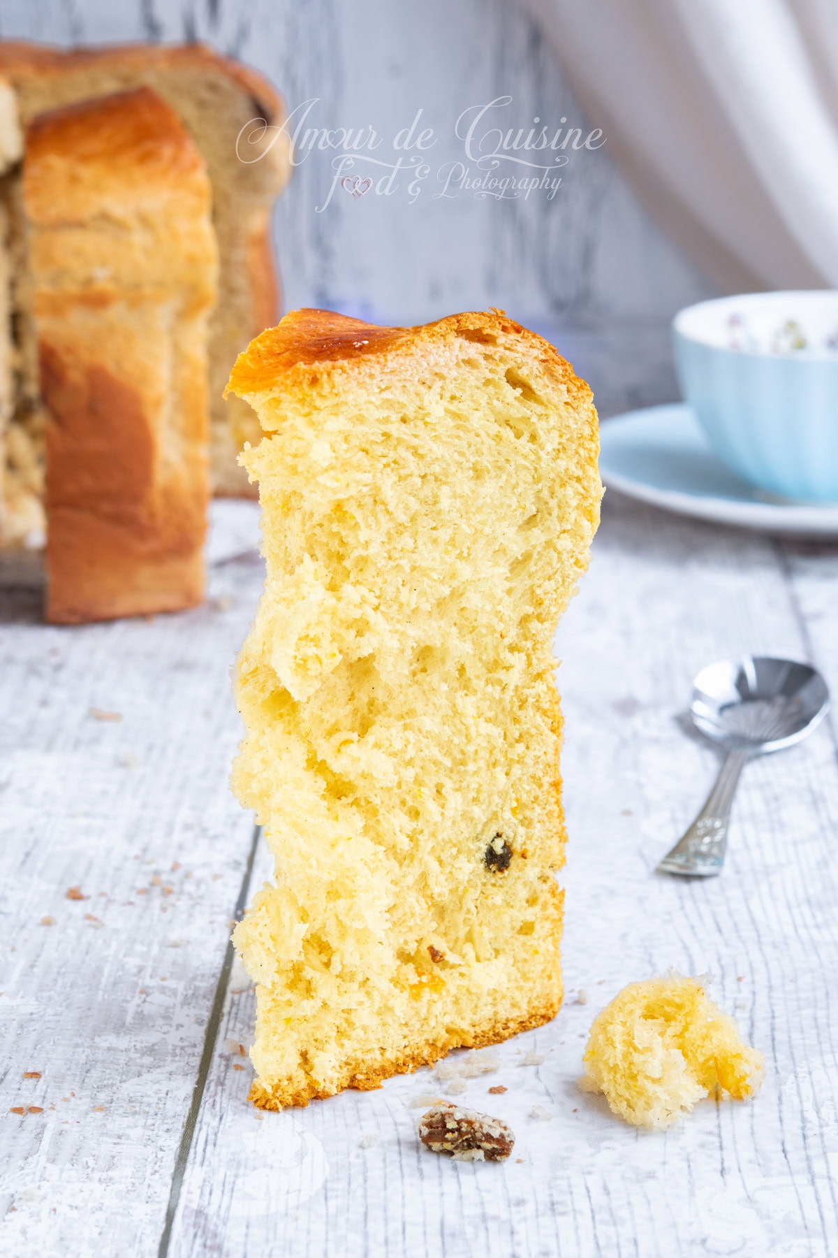 Grande tranche de panettone maison, mie très moelleuse et aérée avec quelques raisins, posée debout sur une table, avec une tasse bleue et le panettone en arrière-plan.