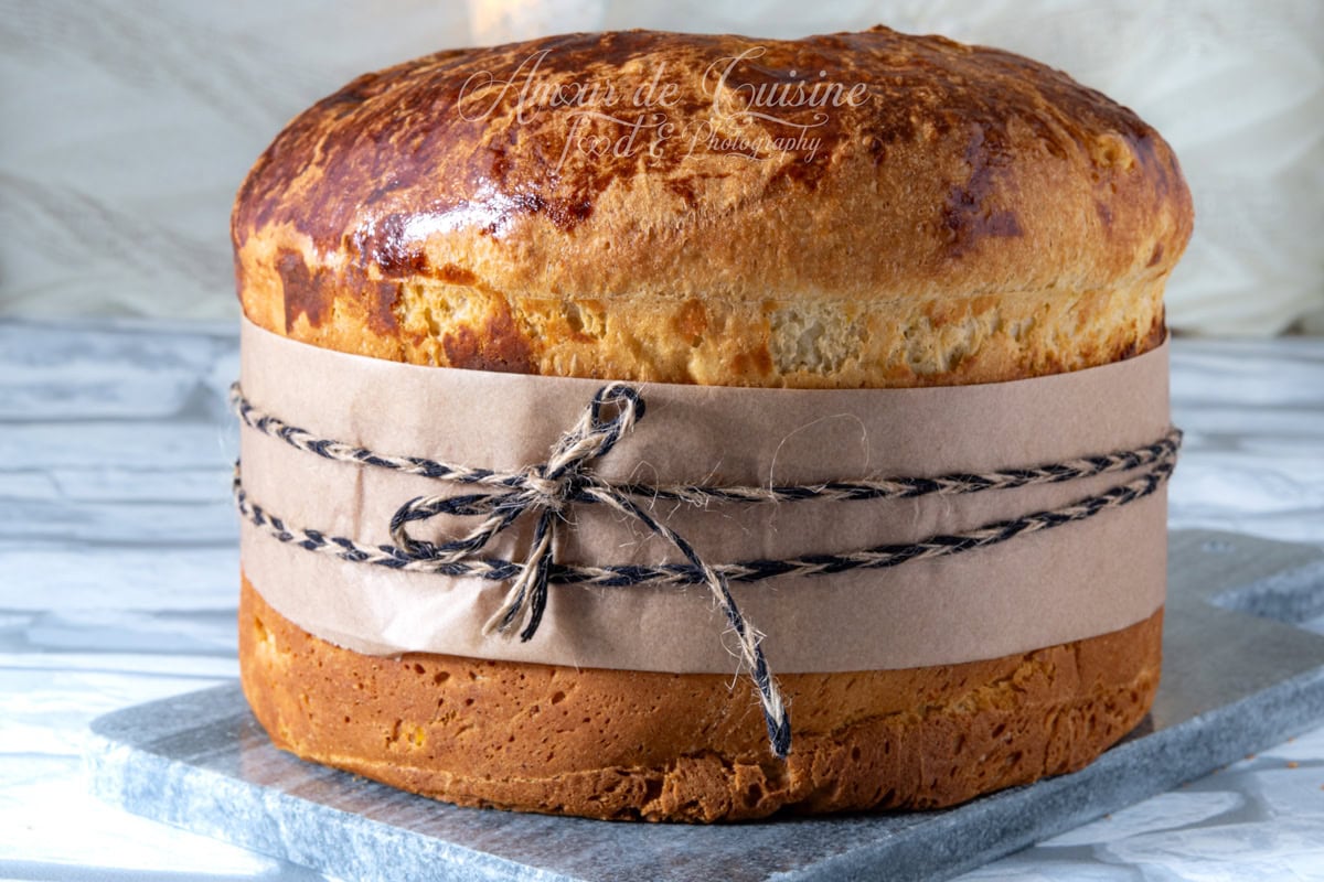 Panettone maison bien doré, présenté entier avec une bande de papier kraft et une ficelle nouée autour, posé sur une planche grise sur fond clair.