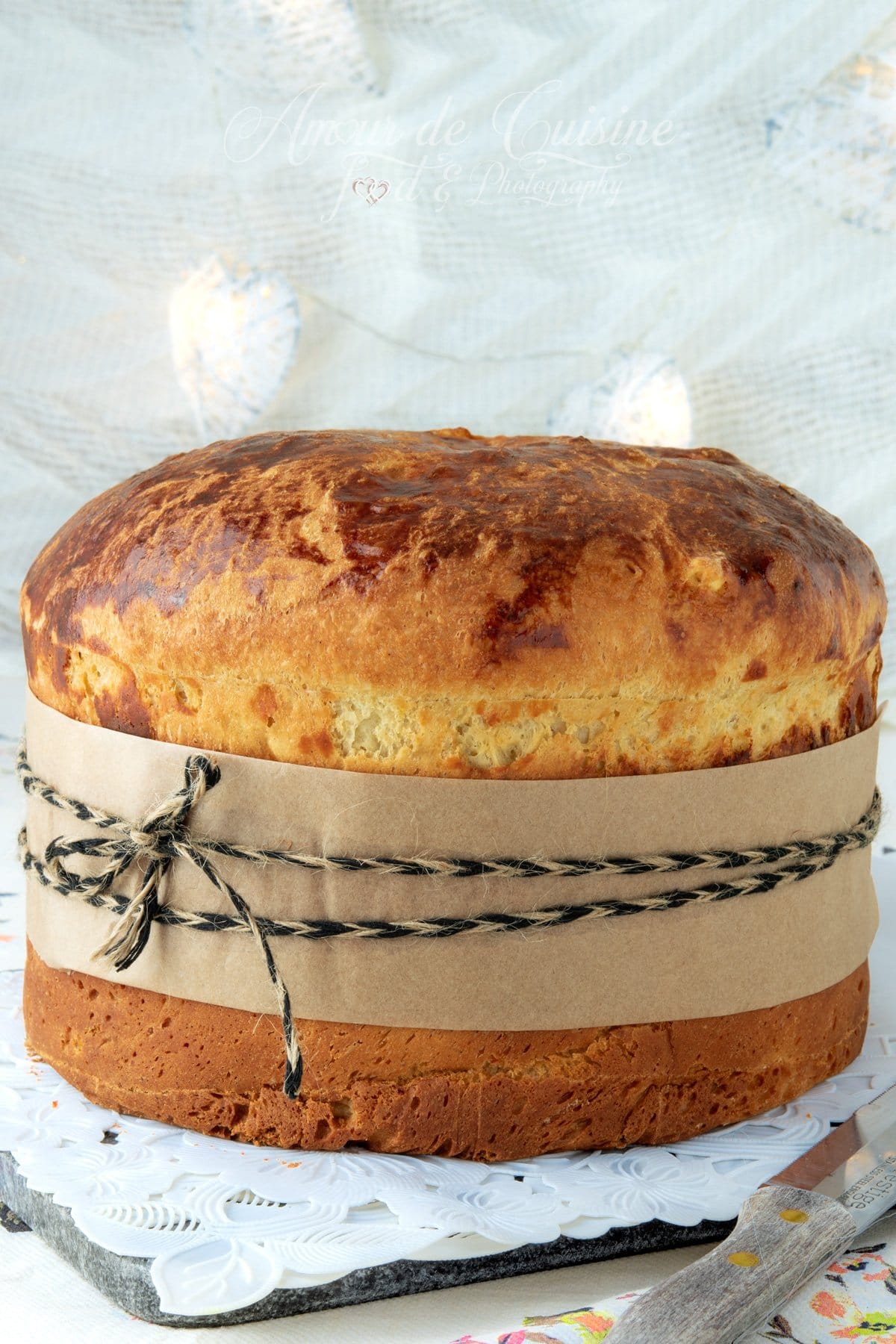 Panettone maison bien doré, haut et moelleux, entouré d’une bande de papier kraft et ficelé, posé sur un napperon blanc.